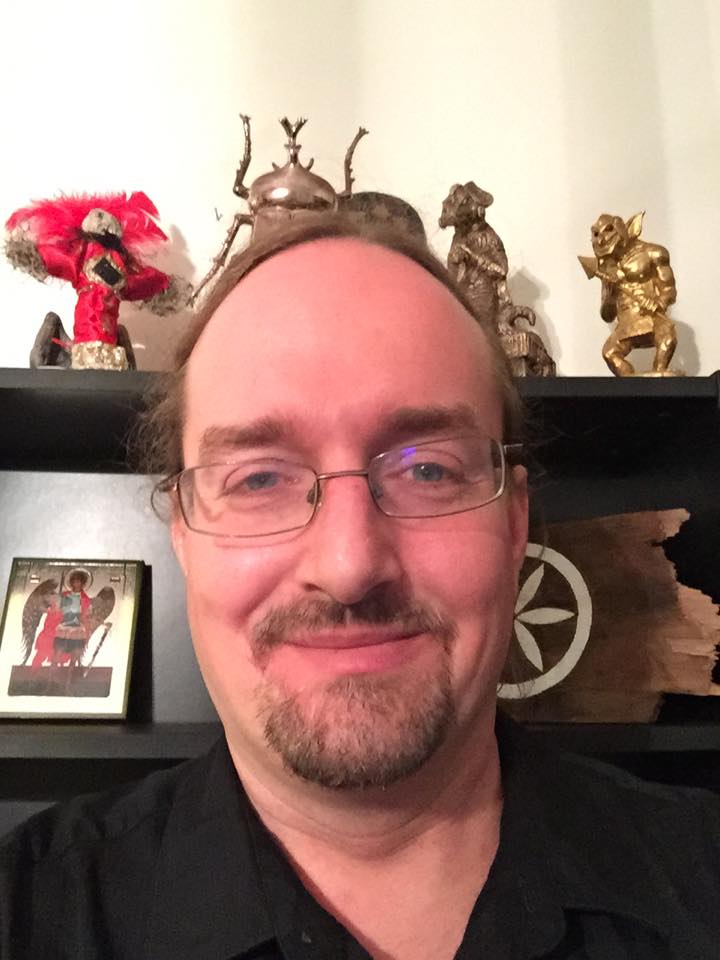 Image is of a Caucasian man with glasses in front of a bookshelf adorned with statuettes.