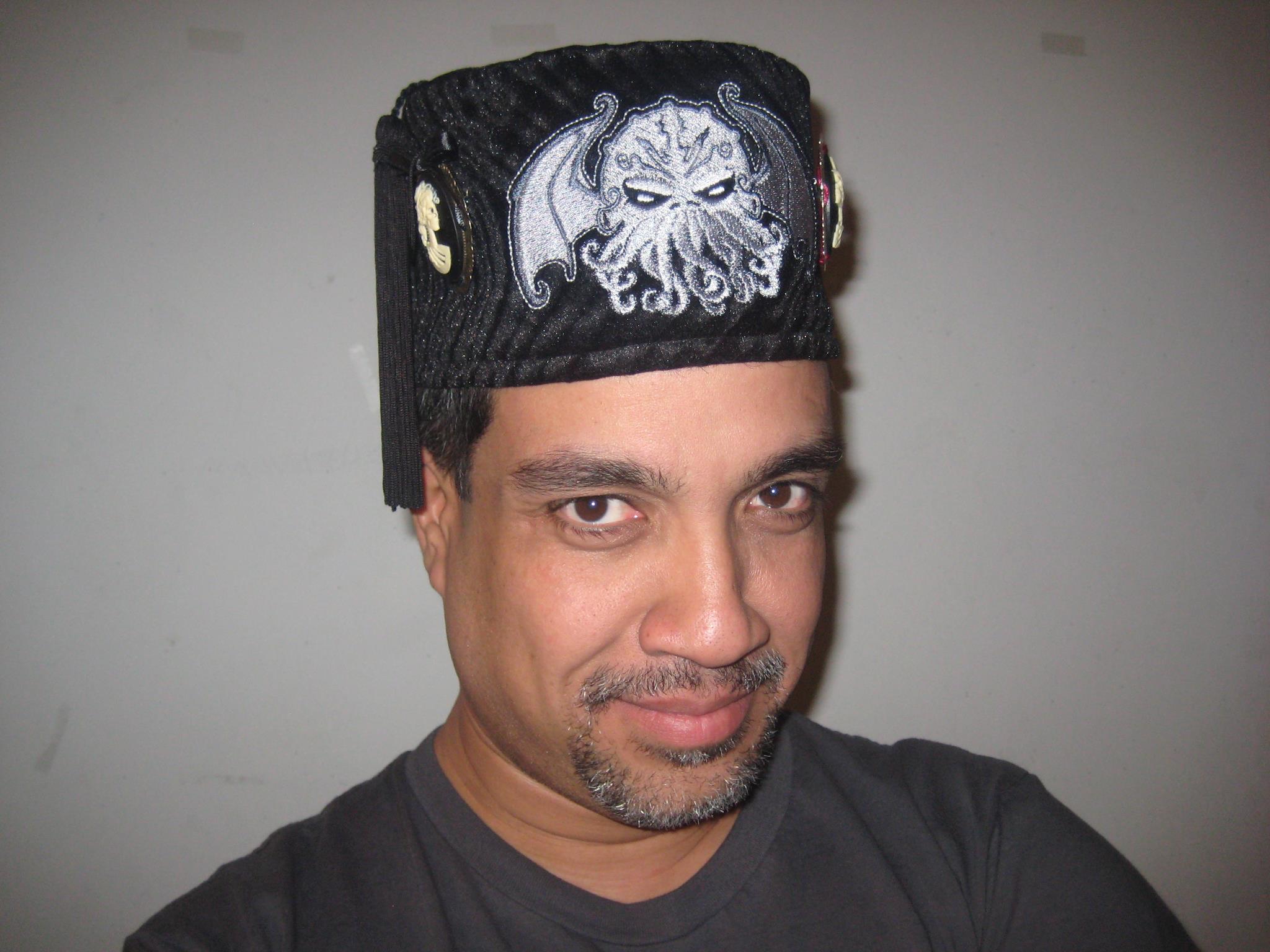 Image is Oscar Rios, a middle aged latino man with a van dyke beard & a black fez bearing the image of cthulhu on it.